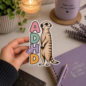 Könnte beinhalten: Ein gestanzter Aufkleber mit einem stehenden Erdmännchen neben dem vertikalen Text ADHD, wobei jeder Buchstabe in einer anderen Pastellfarbe wie Rosa, Grün, Lila und Orange dargestellt ist, eingefasst in einen weißen Rand.
