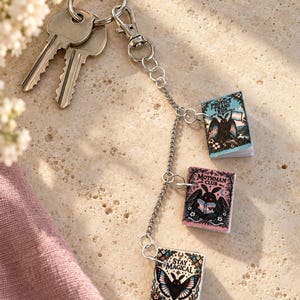 Può includere: Un portachiavi in metallo argentato con un anello, due chiavi e una catenella a cui sono appesi tre ciondoli a forma di libro in miniatura. Il libro in alto è blu con la scritta Protect Trans Kids, quello centrale è rosa con Mothman Club e quello in basso è bianco con Stay Magical.