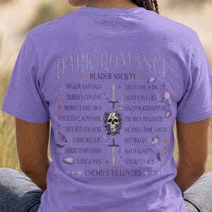 May include: A lilac-colored t-shirt with the text "DARK ROMANCE READER SOCIETY" and a list of related themes. The design includes a skull, swords, and other graphics. The person is sitting on a stone surface.