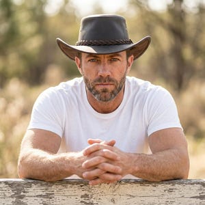 May include: A dark brown leather cowboy hat featuring a braided leather hatband around the crown. The hat has a wide, slightly curved brim and a structured top. The material appears smooth with visible stitching details along the edge of the brim.