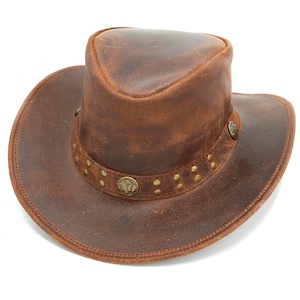 May include: A brown leather cowboy hat with a distressed finish, featuring a wide brim and a pinched crown. The hat is accented with a leather band decorated with small brass studs and circular metal medallions depicting a buffalo profile.