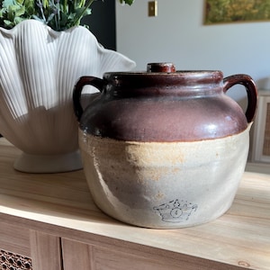 Op de afbeelding: Een aardewerken pot met twee oren, voorzien van een donkerbruin geglazuurde bovenkant en een lichtbeige onderkant. Het vat heeft een bijpassend deksel en een klein, gestempeld kroonembleem aan de voorzijde. De pot heeft een ronde vorm.