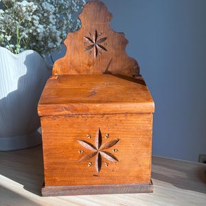 Puede incluir: Una caja de sal de madera de color marrón medio para montar en la pared, con tapa abatible y un motivo floral tallado de seis pétalos tanto en el panel frontal como en el respaldo arqueado. La veta de la madera es visible en todo el recipiente.