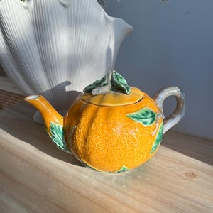 May include: A ceramic teapot shaped like a textured orange fruit with a bright orange body, green leaves, and a brown branch-like handle. The lid features a sculpted green leaf and stem detail, while the spout extends from the side of the fruit.