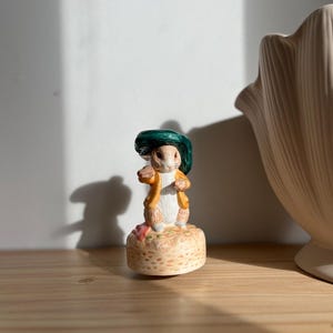 Puede incluir: Figura de cerámica de un conejo con sombrero verde y chaqueta amarilla. El conejo está sobre una base beige con una flor rosa. Es un objeto decorativo.