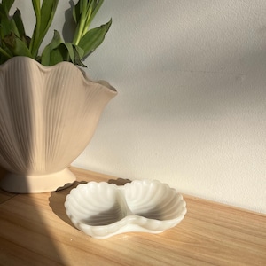 May include: A white, shell-shaped dish with two compartments rests on a wooden surface. The dish has a scalloped edge and a textured, ridged interior. A large, pale pink, shell-shaped vase with green leaves is in the background.