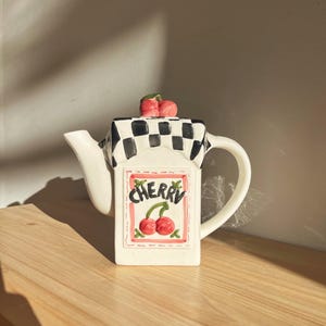 May include: A white ceramic teapot with a black and white checkered pattern on the lid. The teapot features a rectangular label with the word "Cherry" and a cherry illustration. A pair of red cherries tops the lid.