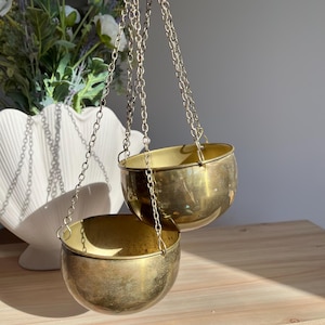 May include: Two vintage brass hanging planters suspended by silver chains. The planters have a rounded bowl shape and a warm, golden hue. The chains are attached to a ring at the top, allowing for easy hanging. The planters are empty and ready for plants.