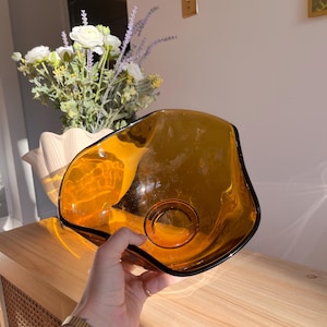 May include: An amber glass bowl with an unusual, irregular shape. The bowl has a dark edge and a circular indentation at the base. The bowl is held over a wooden surface, with a vase of flowers in the background.