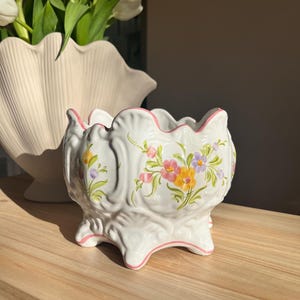 May include: White ceramic planter with a scalloped rim and three feet. The planter is decorated with colorful floral designs in pink, yellow, and purple. A pink trim accents the rim and feet. A second, larger, off-white vase with flowers is in the background.