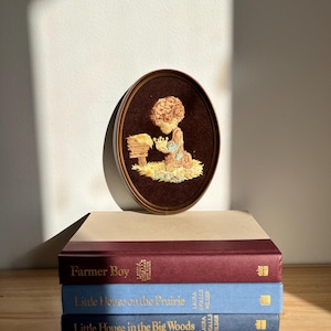 May include: An oval framed embroidery of a child kneeling, with a brown frame and backing. The embroidery features a child with curly hair, wearing a blue and brown outfit. Below the embroidery are three books: "Farmer Boy", "Little House on the Prairie", and "Little House in the Big Woods".