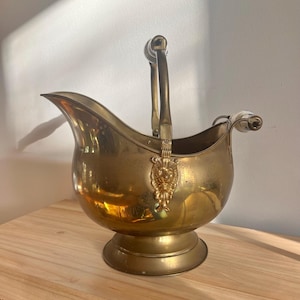May include: A polished brass coal scuttle with a wide pouring spout and a pedestal base. The vessel features a curved handle with a white ceramic grip and decorative lion head mounts where the handle attaches to the main body of the container.