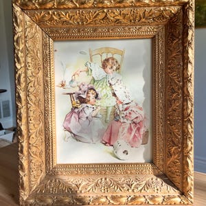 May include: A rectangular gold-toned frame with an ornate, embossed leaf and vine pattern surrounding a print of three children in vintage clothing having a tea party. The children are depicted in soft pastel colors, seated around a small table.