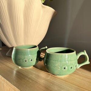 Könnte beinhalten: Zwei kleine, grüne Keramikbecher mit Henkeln und floralen Mustern. Sie stehen auf einer Holzoberfläche, im Hintergrund eine große, cremefarbene dekorative Vase. Die Becher haben eine Craquelé-Glasur.