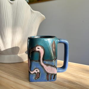 Op de afbeelding: Een vierkante keramische mok met een turquoise glazuur en een flamingo-ontwerp. De roze flamingo's staan tegen een blauwe achtergrond, met bruine poten en snavels. De mok heeft een vierkant handvat.