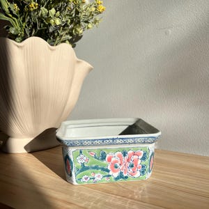 May include: Rectangular ceramic planter with a floral design. The planter features a white base with a green and pink floral pattern. The top edge has a blue and white geometric border. The planter is sitting on a wooden surface.