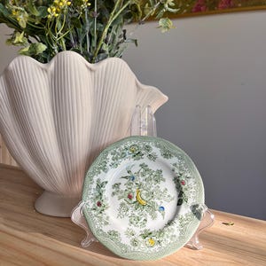 May include: A small round ceramic plate featuring a detailed green floral and bird pattern on a white background. The plate has a decorative green border with intricate scrollwork and is displayed on a clear plastic stand for viewing.
