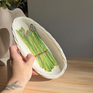 May include: White ceramic asparagus serving dish with a woven rim. The dish features a realistic depiction of green asparagus spears tied with a brown twine. A hand is holding the dish on a wooden surface.