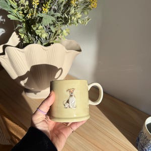Puede incluir: Una taza de cerámica amarillo claro con asa, con una ilustración de perro. La taza se sostiene en una mano, con un arreglo floral en un jarrón beige en el fondo. La taza es de tamaño estándar.