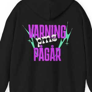 May include: A black hooded sweatshirt featuring a graphic on the back with the text VARNING pms PÅGÅR in purple and white lettering. The design includes teal starburst accents and is centered on the back of the garment, which has long sleeves.