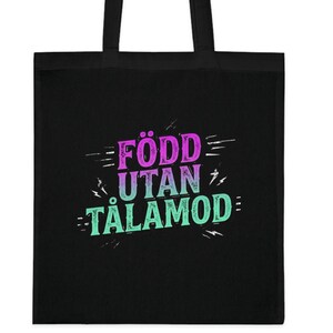 May include: Black tote bag with the text "FÖDD UTAN TÅLAMOD" in purple, teal, and white. The tote bag has black handles and is made of a durable fabric. The text is in a stylized font.