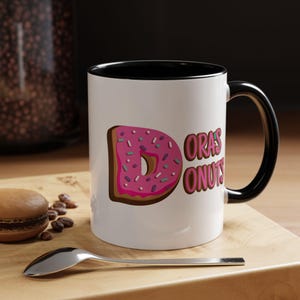 May include: White ceramic mug with a black interior and handle. The mug features a pink donut graphic with sprinkles and the text "Dora's Donuts" in pink. A spoon, a macaron, and coffee beans are on a wooden surface.