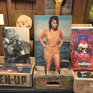 Puede incluir: Una exhibición de grabados artísticos vintage, que incluye una imagen en blanco y negro de un músico tocando la trompeta, una impresión en color de una mujer de pie en el agua y un cartel de concierto psicodélico. Los grabados se exhiben en cajas de madera.