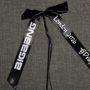 May include: A black satin ribbon tied into a bow with two long tails. The left tail features the white text BIGBANG, while the right tail displays the white text London 2026 and GDragon. The ribbon has a smooth, reflective finish.