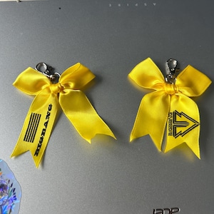 May include: Two bright yellow satin ribbon bows, each attached to a silver-toned metal lobster clasp. The left bow features the text BIGBANG and five vertical lines in black ink. The right bow displays a black triangular logo and the text BIGBANG.