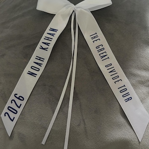 May include: White satin ribbon bow with navy blue text. The text reads "NOAH KAHAN 2026" on one side and "THE GREAT DIVIDE TOUR" on the other. The bow has long, thin streamers.