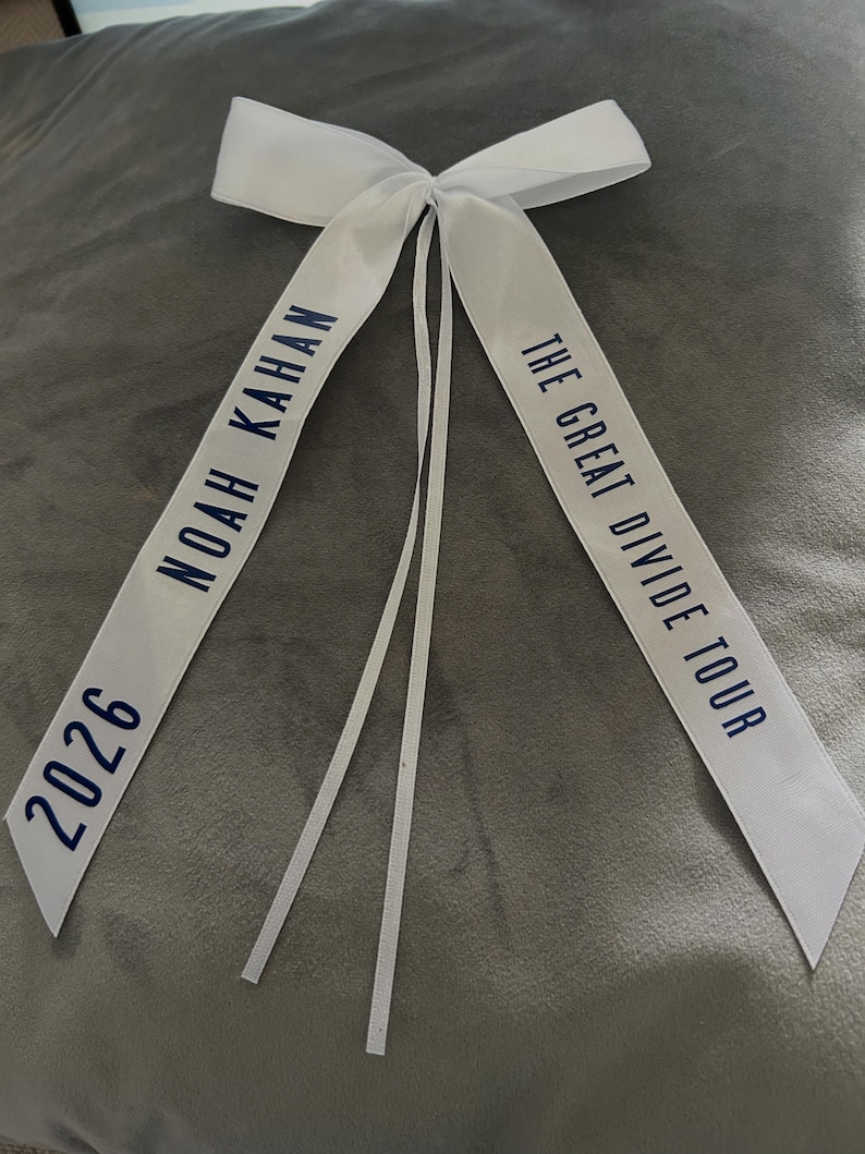 May include: White satin ribbon bow with navy blue text. The text reads "NOAH KAHAN 2026" on one side and "THE GREAT DIVIDE TOUR" on the other. The bow has long, thin streamers.