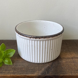May include: A round, white ceramic ramekin featuring a ribbed exterior texture and a dark brown, speckled rim. The bowl has a speckled finish throughout its surface and is accompanied by a small sprig of fresh green basil leaves placed beside it.