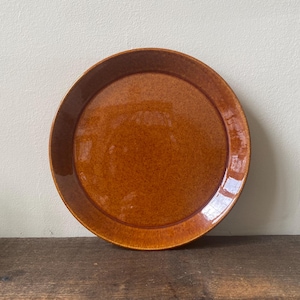 May include: A round, brown ceramic plate with a speckled glaze. The plate has a slightly raised rim and a glossy finish. The plate is sitting on a wooden surface. The plate is approximately 25 cm in diameter.