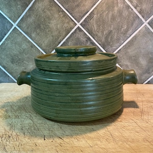 May include: A round, olive green ceramic casserole dish with a matching lid. The pot features horizontal ribbed detailing along the exterior walls and two small, integrated side handles. The lid has a flat top and a circular knob handle.