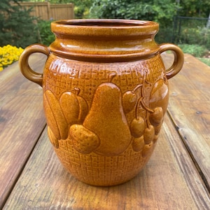 May include: A glazed amber-coloured ceramic jar featuring two side handles and a raised relief pattern of various fruits, including a prominent pear and grapes, set against a textured grid background on the exterior surface.