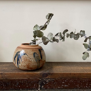 May include: A round, speckled tan ceramic vase with a short neck and a wide body, featuring dark blue glaze drips running down the sides. A single dried eucalyptus branch with multiple round, muted green leaves extends from the opening.