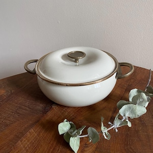 May include: A round, off-white enamelled casserole dish with a matching lid, featuring a metallic brass-toned rim, two side handles, and a central knob on the lid. The pot has a smooth, glossy finish and a wide, shallow bowl-like shape.