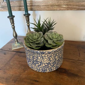 May include: A round, blue and white ceramic pot with a textured pattern, filled with green succulents and other plants. The pot is on a wooden surface. Two brass candle holders with green candles are in the background.