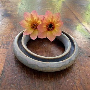 May include: A circular, light blue ceramic ring with a smooth, glazed finish. Two peach and yellow dahlia flowers with dark centres are resting on the top edge of the ceramic piece, partially overlapping the inner opening of the ring.