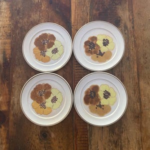 May include: A set of four round white ceramic plates, each featuring a central floral motif composed of three overlapping flowers in shades of brown, tan, and pale yellow. Each plate has a thin metallic gold rim along the outer edge.
