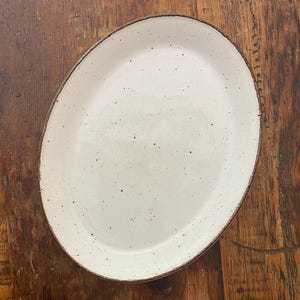 Peut inclure: Un plat ovale en céramique doté d'un vernis blanc cassé moucheté et d'un bord brun foncé. La surface de l'assiette est lisse avec des taches sombres visibles réparties sur la base claire, créant un contraste marqué avec le contour du plat.
