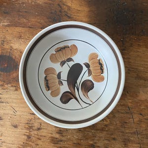 May include: A round ceramic bowl with a cream-coloured base and a dark brown rim. The centre of the bowl features a painted floral design with three tan flowers and dark brown leaves, all enclosed within a thin dark brown circular line.