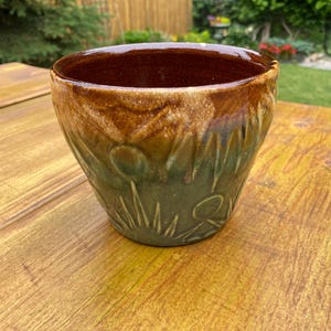 May include: A ceramic cup with a wide, circular opening and a tapered base. The interior is finished in a glossy dark brown glaze, while the exterior features a textured, earthy green glaze with carved sun and mountain motifs near the bottom.