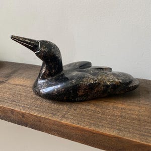 Peut inclure: Une sculpture en pierre sombre polie représentant un plongeon, avec des textures mouchetées de gris et de noir. La figurine présente un long bec pointu avec une ligne blanche gravée près de la base et une signature inscrite en blanc sur le côté.