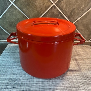 May include: A vibrant red cooking pot with a matching lid. The pot features two side handles and a handle on the lid. It is placed on a grey patterned surface.