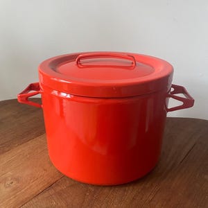 Peut inclure: Une marmite en métal émaillé orange vif avec un couvercle assorti et deux poignées latérales. La marmite cylindrique présente une finition lisse et brillante ainsi qu'un couvercle plat avec une poignée centrale, offrant un aspect uniforme.