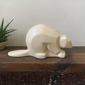 May include: A smooth, cream-coloured ceramic figurine shaped like a beaver in a stylised, geometric pose. The sculpture features a rounded back, a flat tail extending horizontally, and a simplified head profile, all finished with a glossy, reflective surface.