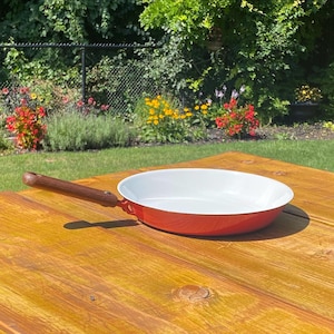 Peut inclure: Une poêle ronde avec un extérieur orange vif et un revêtement intérieur blanc lisse. La poêle est dotée d'un long manche droit en bois fixé par un joint métallique, créant un contraste entre le bois chaud et la couleur orange vive.