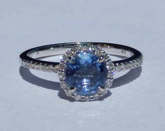 Op de afbeelding: Een zilveren ring met een blauwe edelsteen omringd door een halo van kleinere diamanten. De band is ook bezet met kleinere diamanten.