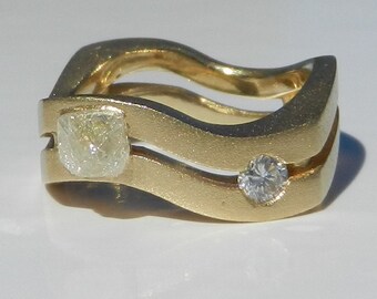 May include: A gold ring with two diamonds. The larger diamond is a square shape and the smaller diamond is round. The ring is designed with two bands that are curved and joined together.